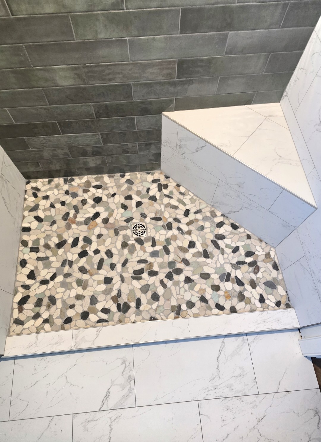 Shower floor with a pebble-style mosaic tile pattern, a central round drain, marble-look tiles on the shower bench and walls, and rectangular gray wall tiles in the background.