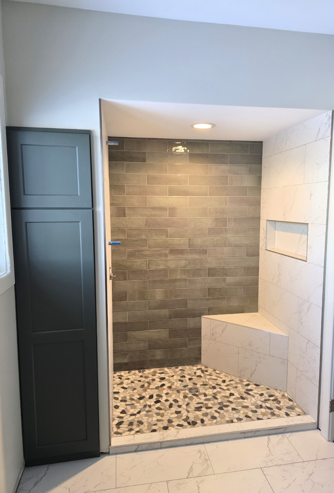 A modern shower with gray subway tiles on the back wall, a built-in bench and shelf, and a pebble-style tile floor. The surrounding walls and floor are white with subtle gray veining. A gray cabinet is to the left.