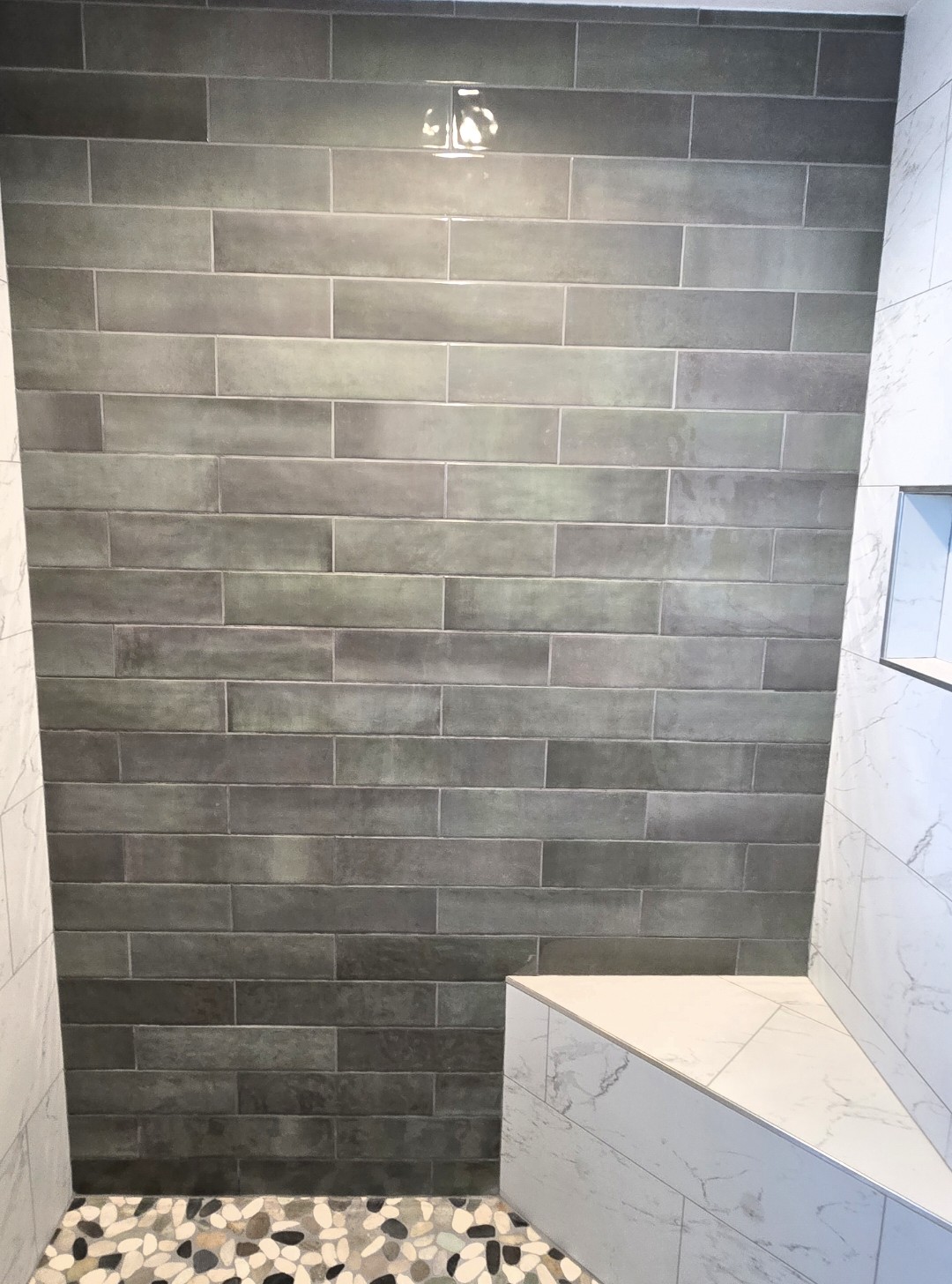 A shower with a dark gray subway tile accent wall, white marble-look tiles on the side, a built-in bench, a recessed shelf, and a pebble-style floor.