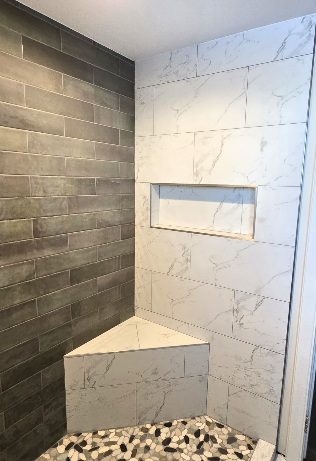 Modern shower with white marble-look wall tiles, a built-in niche shelf, a corner bench, dark green subway tiles on one wall, and a floor with black, white, and gray pebble-shaped tiles.