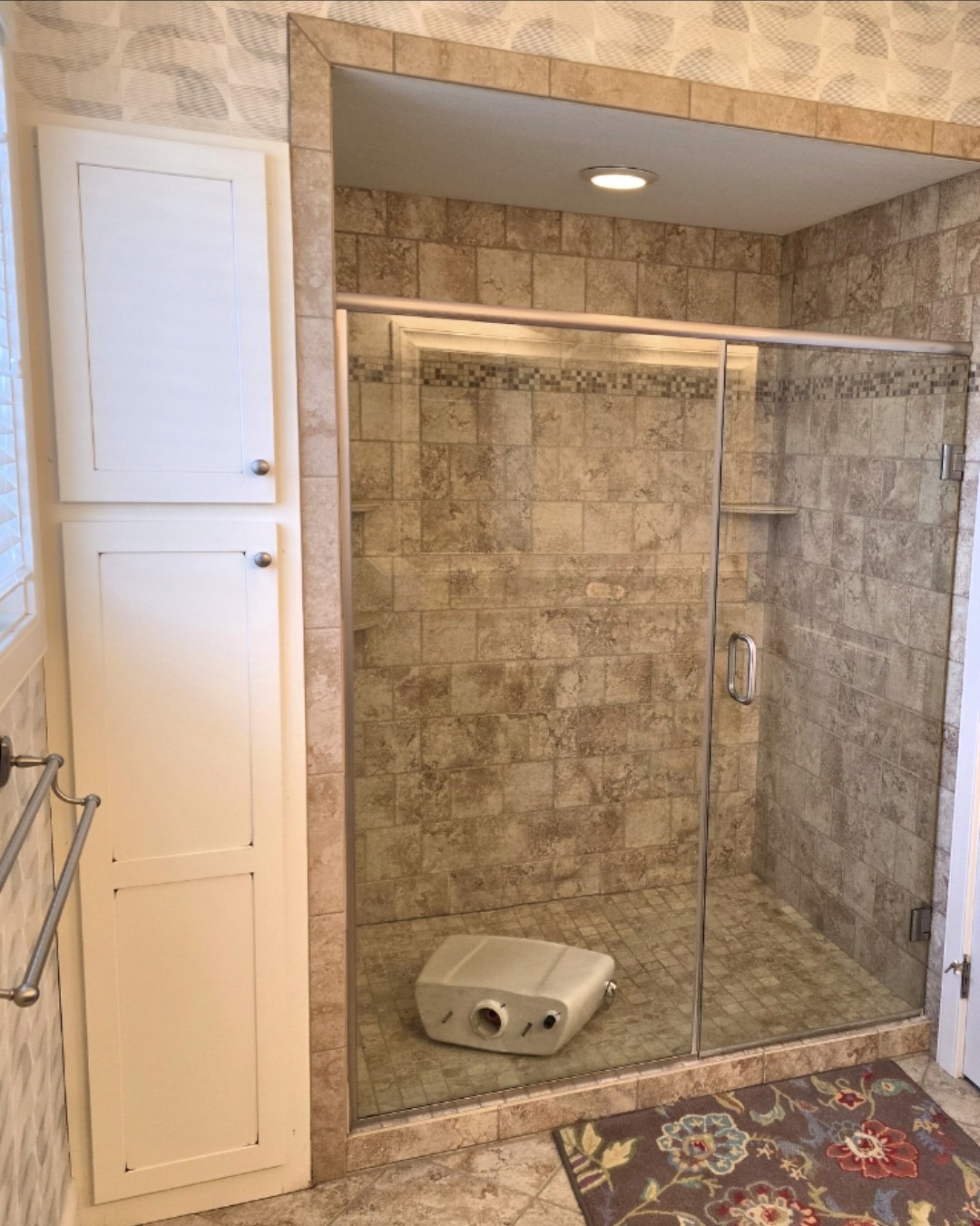 A tiled walk-in shower with a glass door, built-in shelf, and beige walls. A plastic shower chair is lying on its side inside. A floral rug and white cabinets are visible outside the shower.