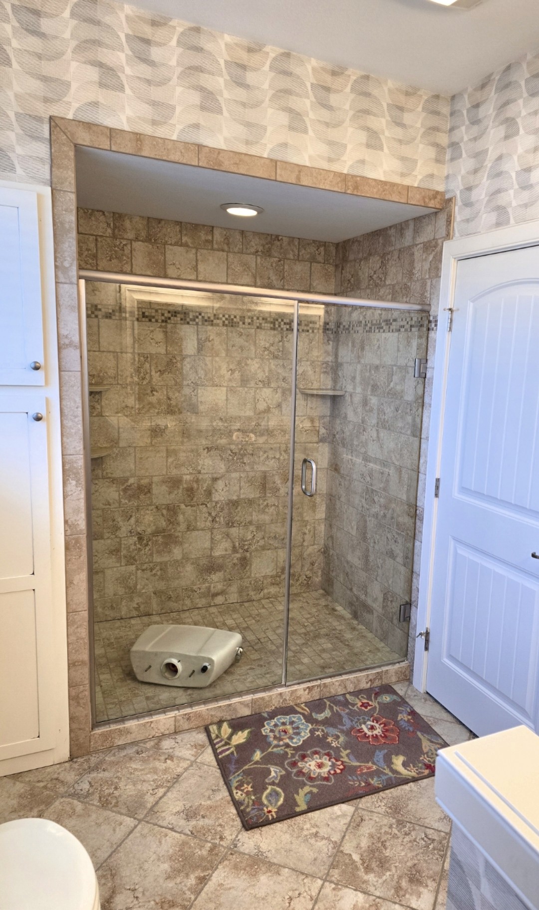 A tiled shower with a glass door, a brown and beige mosaic tile interior, and a small shelf. A white shower seat is on the floor inside, and a floral rug lies in front of the shower on the tile floor.